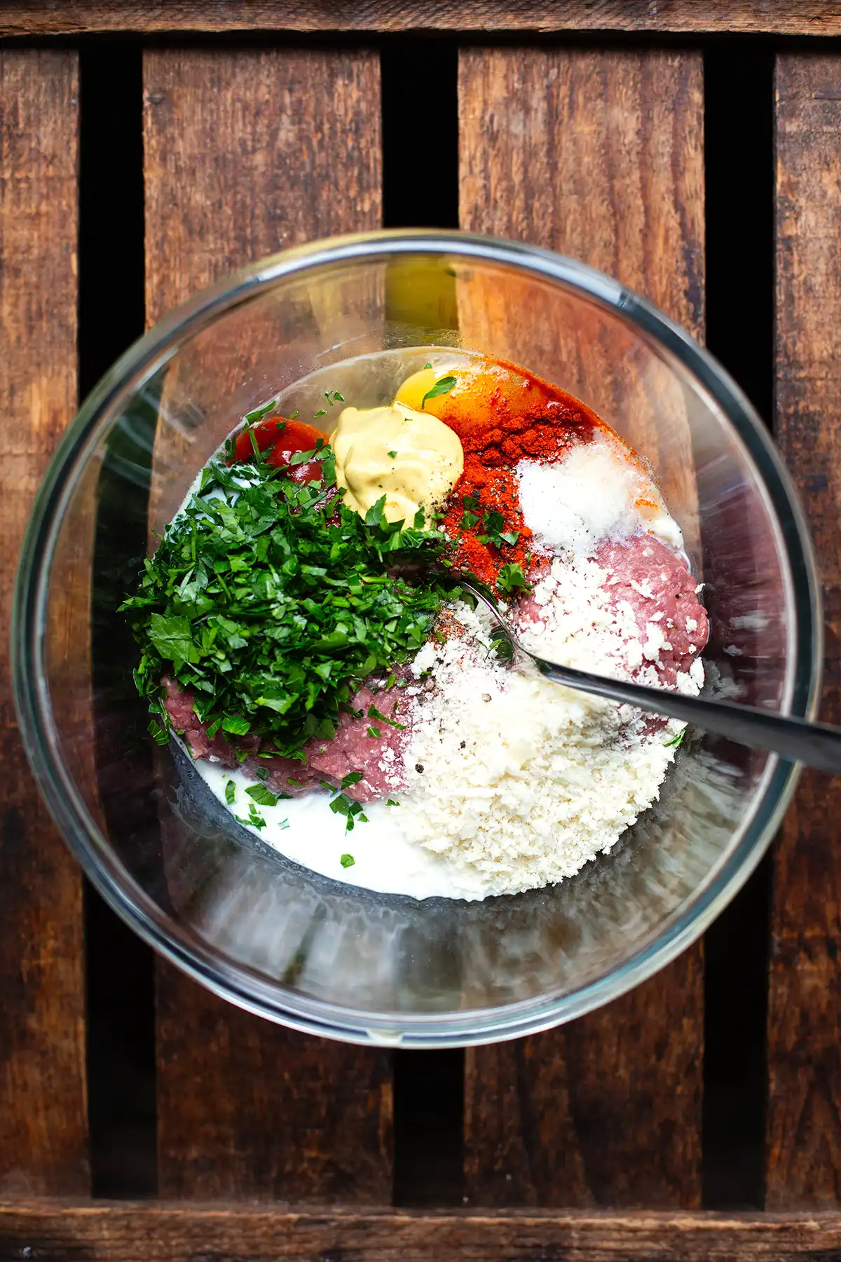 Alle Zutaten für saftige Frikadellen wie von Oma in einer Glasschüssel mit einem silbernen Löffel - bereit zum Umrühren. Diese steht auf einem dunklen Holzuntergrund.