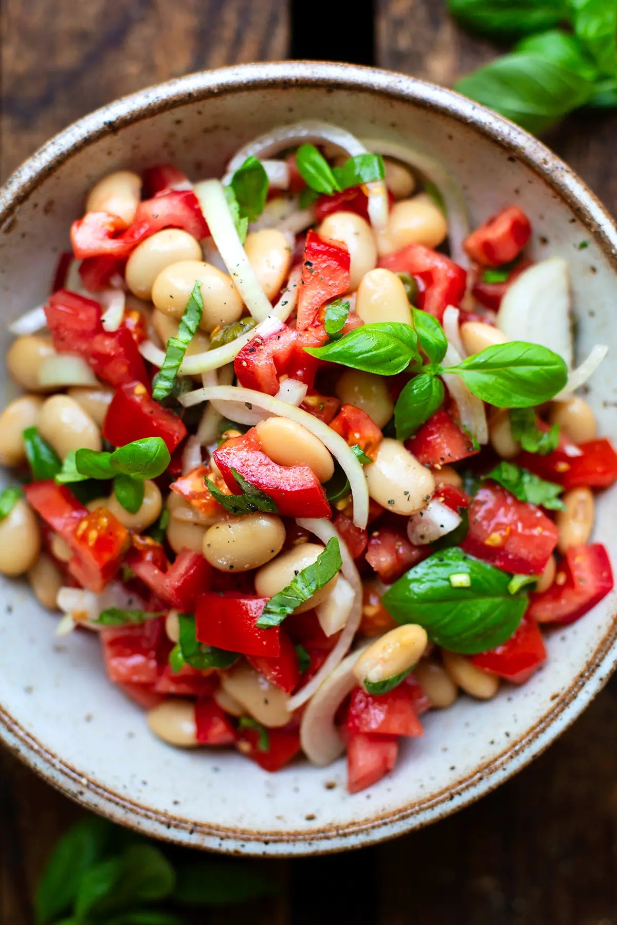 Fertiger weiße Bohnensalat in einer grau melierten Schüssel garniert mit frischem Basilikum. Die Schüssel steht auf einem dunklen Holzuntergrund.