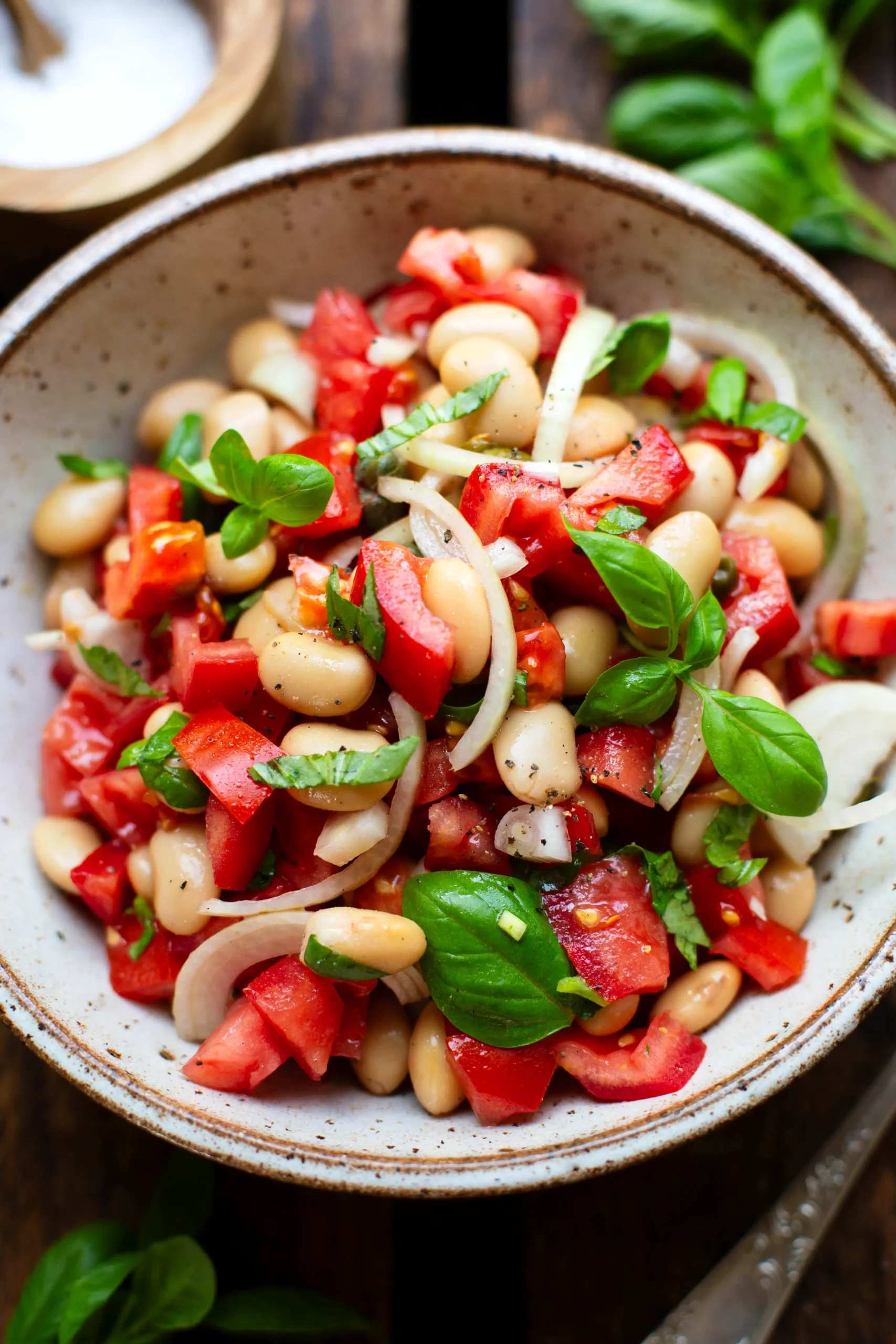 Der fertige weiße Bohnen Salat in einer grau melierten Schüssel. Garniert mit frischem Basilikum. Die Schüssel steht auf einem dunklen Holzuntergrund. Neben der Schüssel zu sehen ist eine silberne Gabel.