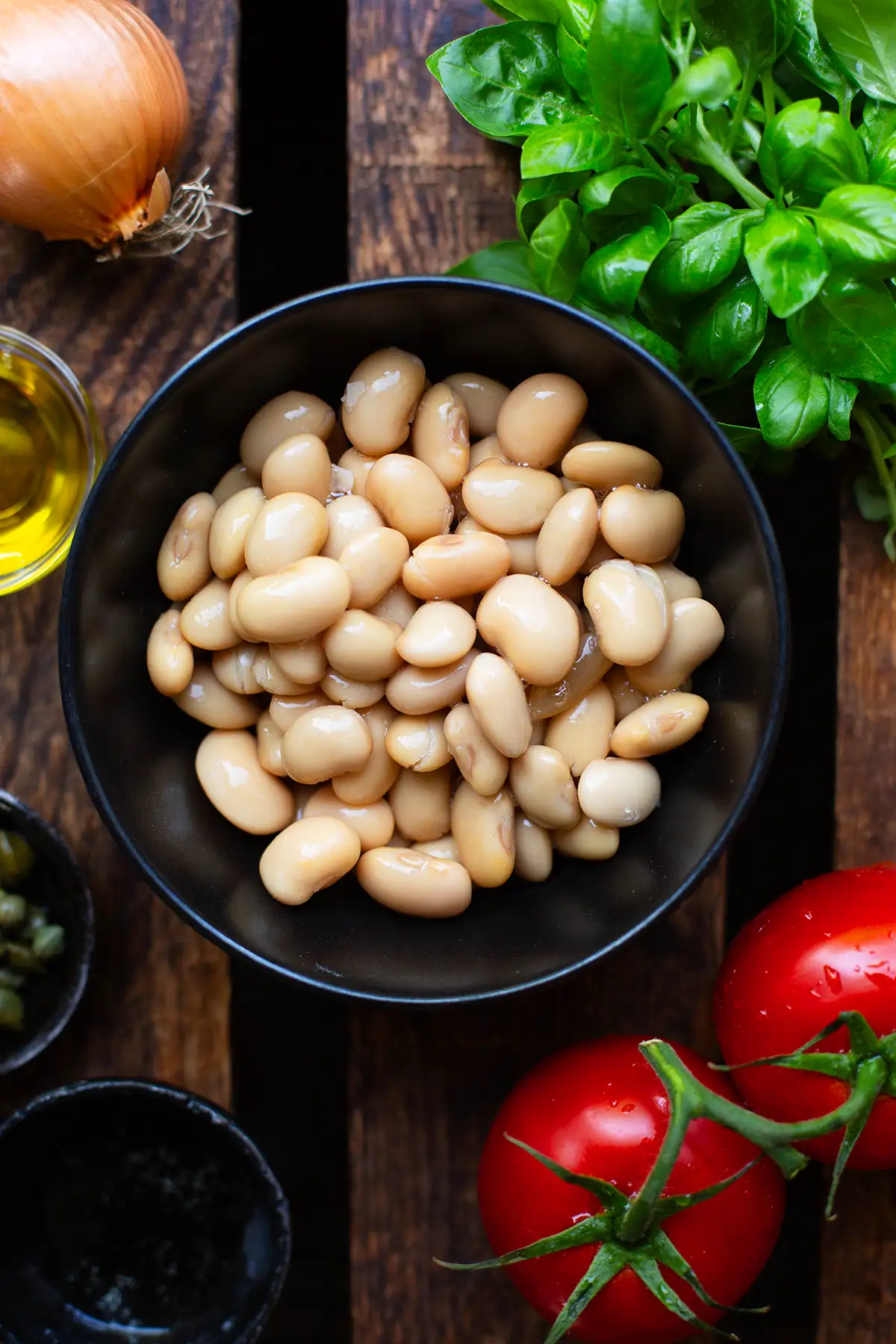 Alle Zutaten für den weiße Bohnen Salat einzeln in Schüsselchen auf einem dunklen Holzuntergrund. Zu sehen sind weiße Riesenbohnen, Olivenöl, Weißweinessig, Basilikum, weiße Zwiebel, Tomaten und Kapern. 