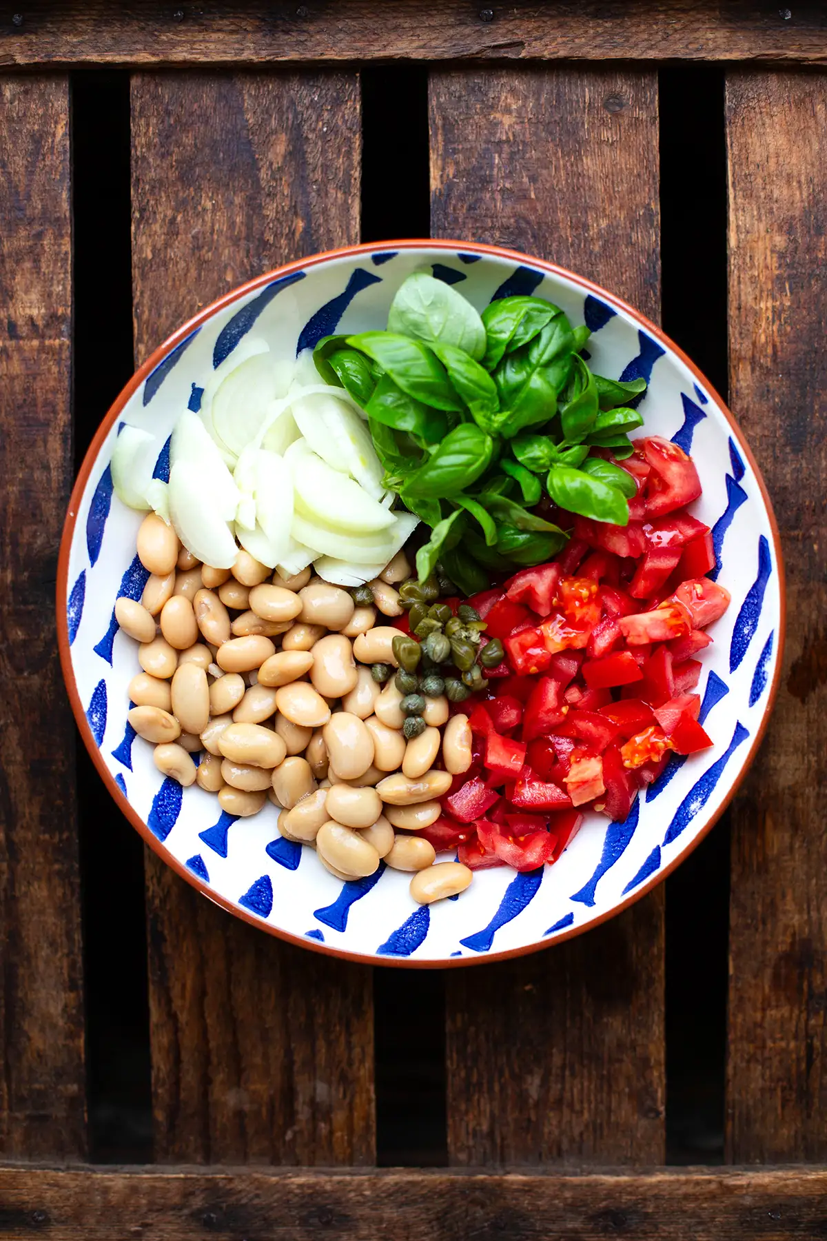 Die festen Zutaten für den weiße Bohnensalat in einer weißen Schüssel mit braunem Rand und blauen Fischen. Zu sehen sind, weiße Bohnen, klein geschnittene Tomaten, Zwiebel, Kapern und frischer Basilikum. Die Schüssel steht auf einem dunklen Holzuntergrund.