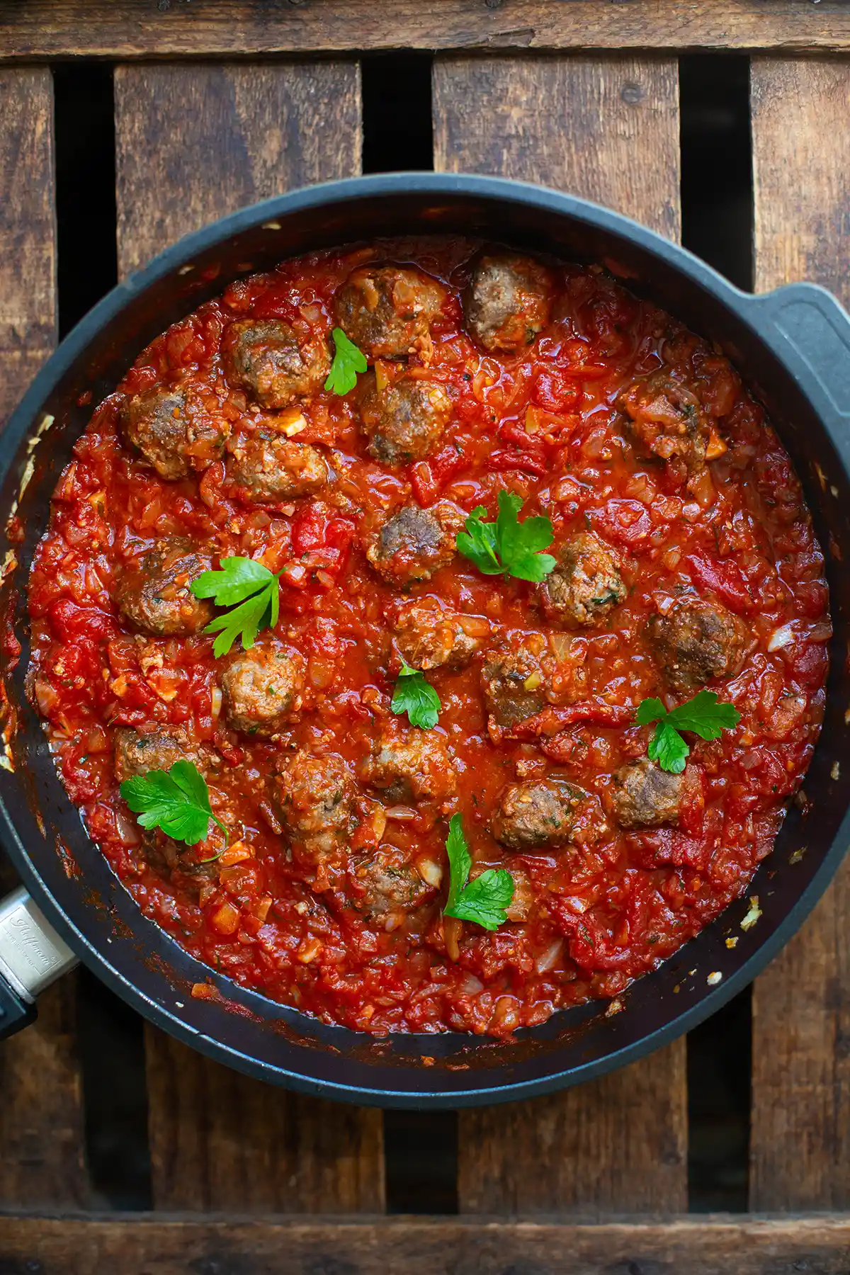 Fertige Hackbällchen in Tomatensauce in einer schwarzen Pfanne, garniert mit glatter Petersilie. Die Pfanne steht auf einem dunklen Holzuntergrund.