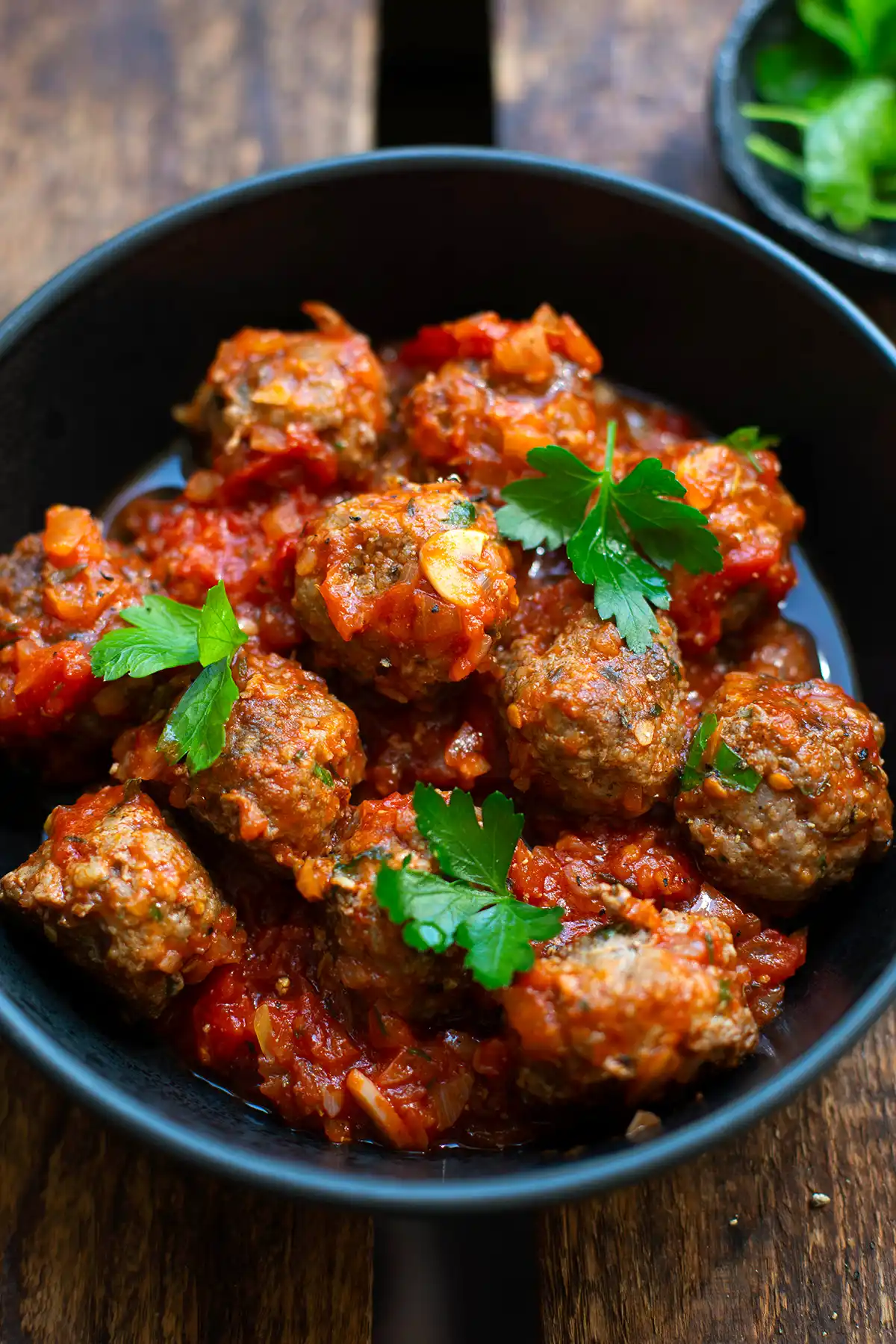 Fertige spanische Hackbällchen mit Tomatensauce auf einem schwarzen Teller, garniert mit glatter Petersilie. Der Teller steht auf einem dunklen Holzuntergrund. Im Hintergrund ist ein kleinen schwarzes Schälchen mit weiterer Petersilie zum Garnieren zu sehen.