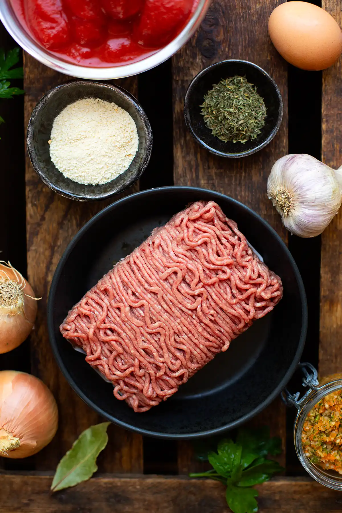 Die Zutaten für spanische Albondigas einzeln auf dunklem Holzuntergrund. Zu sehen sind: gemischtes Hackfleisch, Knoblauch, weiße Zwiebeln, geschälte Tomaten aus der Dose, Thymian, Gemüsebrühpaste, Paniermehl und ein Ei, 