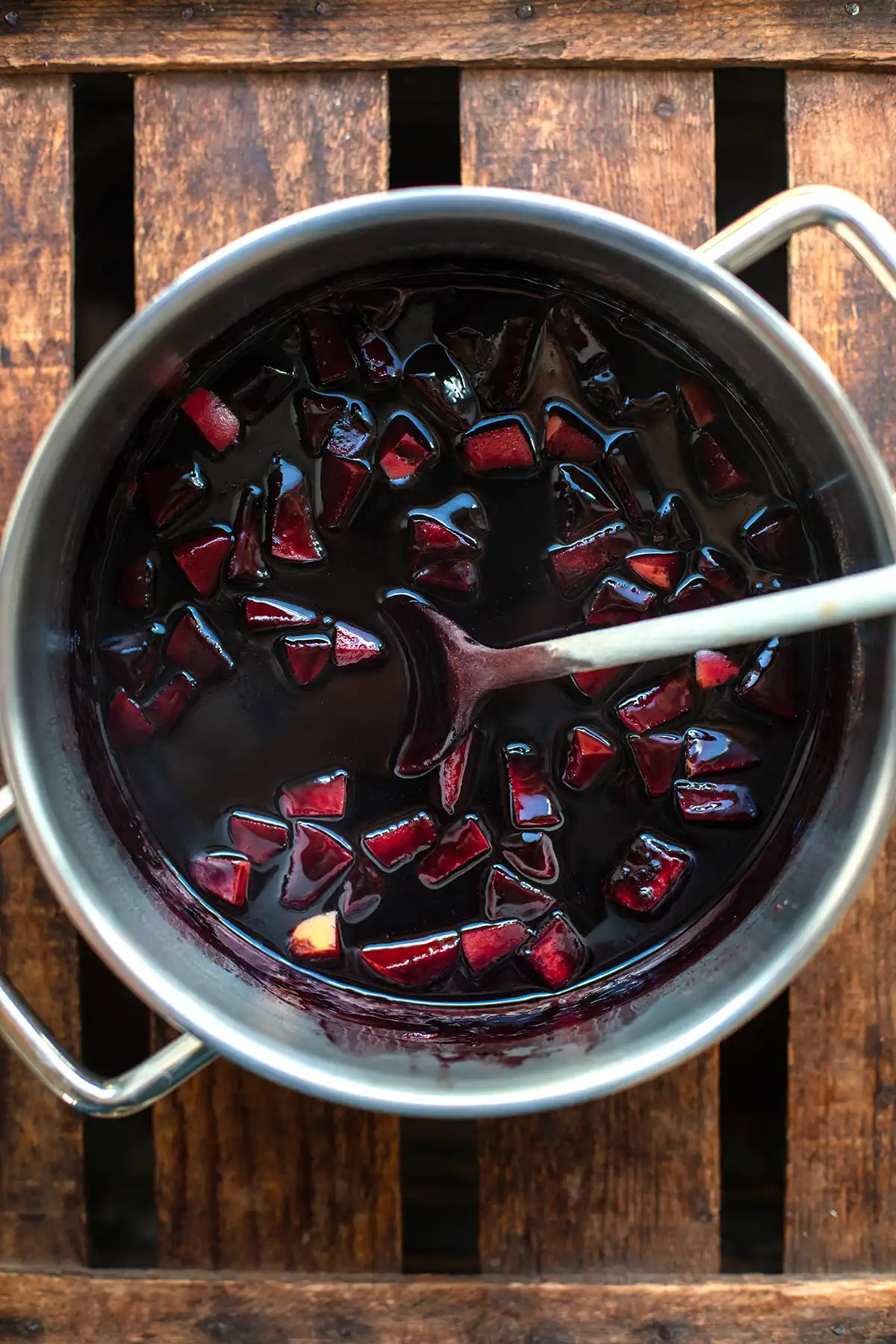 Norddeutsche Fliederbeersuppe in einem silbernen Topf mit Apfelstückchen. Ein Holzlöffel lehnt im Topf. Der Topf steht auf einem dunklen Holzuntergrund.