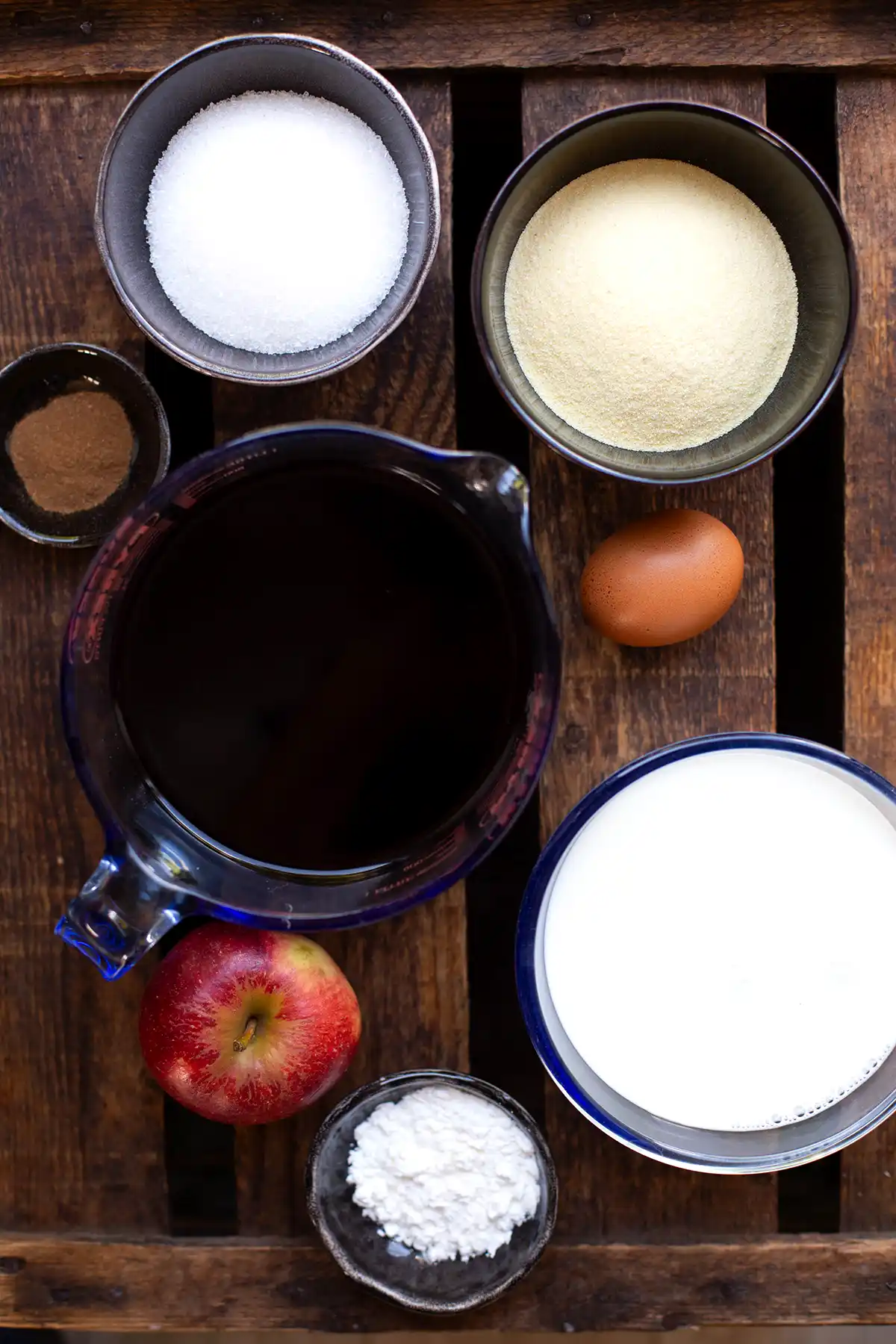 Die Zutaten für Omas Fliederbeersuppe einzeln auf dunklem Holzuntergrund. Zu sehen sind, Speisestärke, Milch, ein ganzer Apfel, Fliederbeersaft, ein Ei, Grieß, Zimt und Zucker.