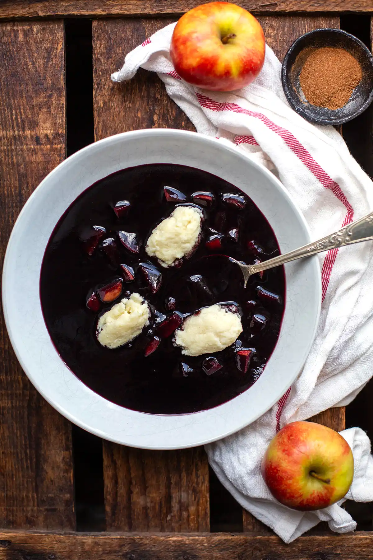 Die fertige Fliedersuppe wie von Oma in einem weißen Suppenteller. Garniert mit drei Grießklößchen und einem silbernen Löffel. Neben dem Suppenteller liegt ein rot-weißes Geschirrtuch, Zimt in einer kleinen Schüssel und zwei ganze Äpfel.
