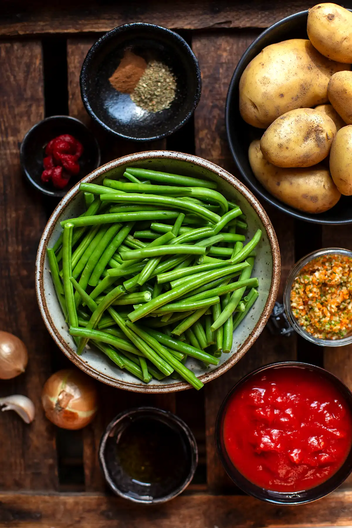Zutaten für den Fasolakia Grüne Bohnen Eintopf in einzelnen Schüsselchen auf dunklem Holzuntergrund.
Zu sehen sind Kartoffeln, Gewürze, Tomatenmark, grüne Bohnen, selbstgemachte Gemüsebrüh-Paste, weiße Zwiebeln, gehackte Tomaten und Öl.