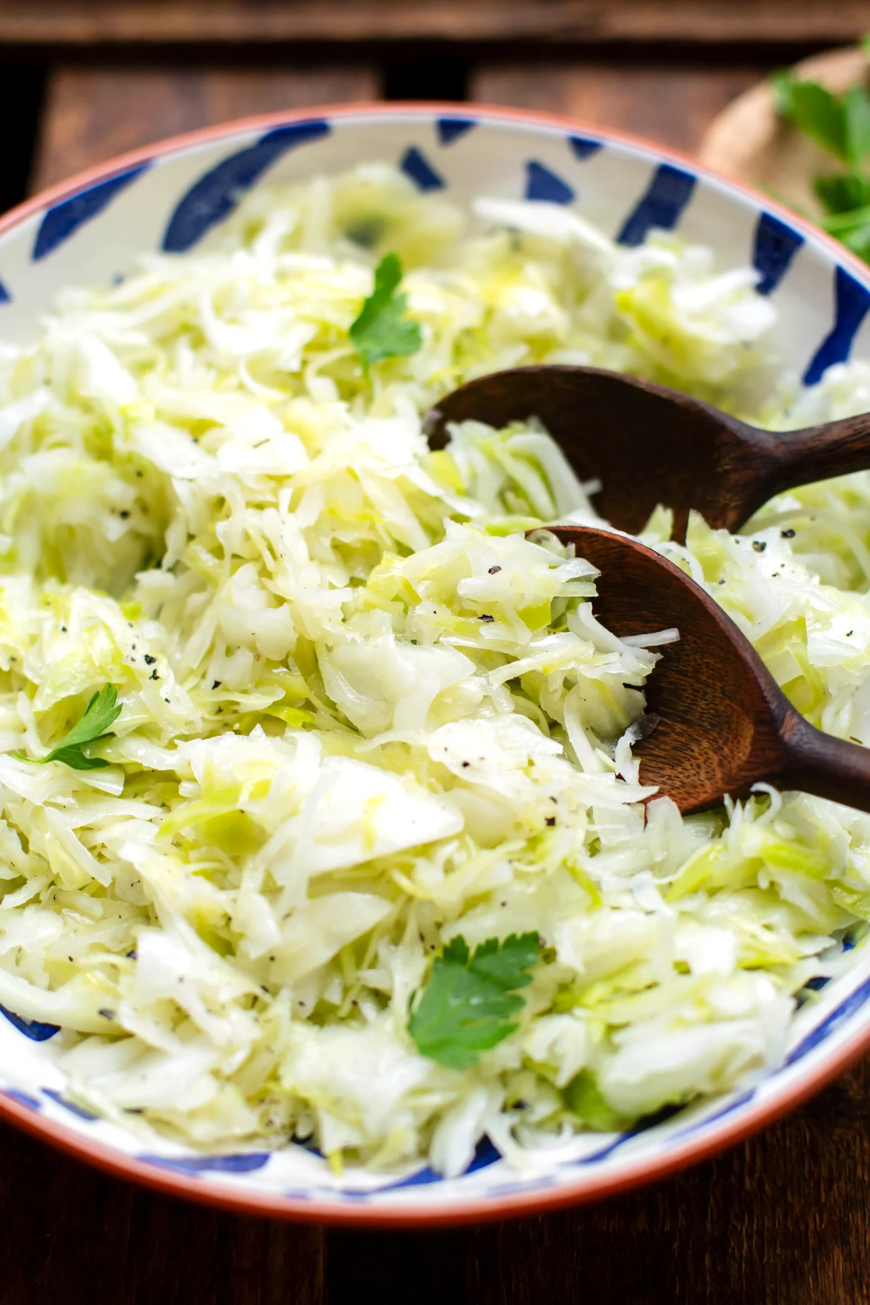 Fertiger griechischer Krautsalat in einer großen Schüssel, garniert mit glatter Petersilie. Im griechischen Krautsalat stecken zwei große Holzlöffel - bereit zum Servieren. Die Schüssel steht auf einem dunklen Holzuntergrund und im Hintergrund ist weitere Petersilie zum Garnieren zu sehen.