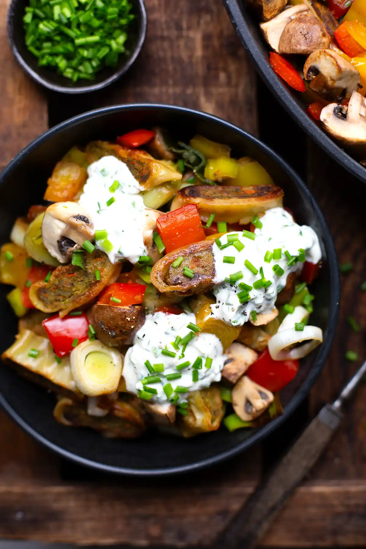 Fertige vegetarische Maultaschen Pfanne aufgeteilt auf zwei schwarze Schüsseln.. Garniert mit Schnittlauch-Schmand. Daneben steht eine kleine Schüssel mit gehacktem Schnittlauch. Die Schüsseln stehen auf einem dunklem Holzuntergrund.