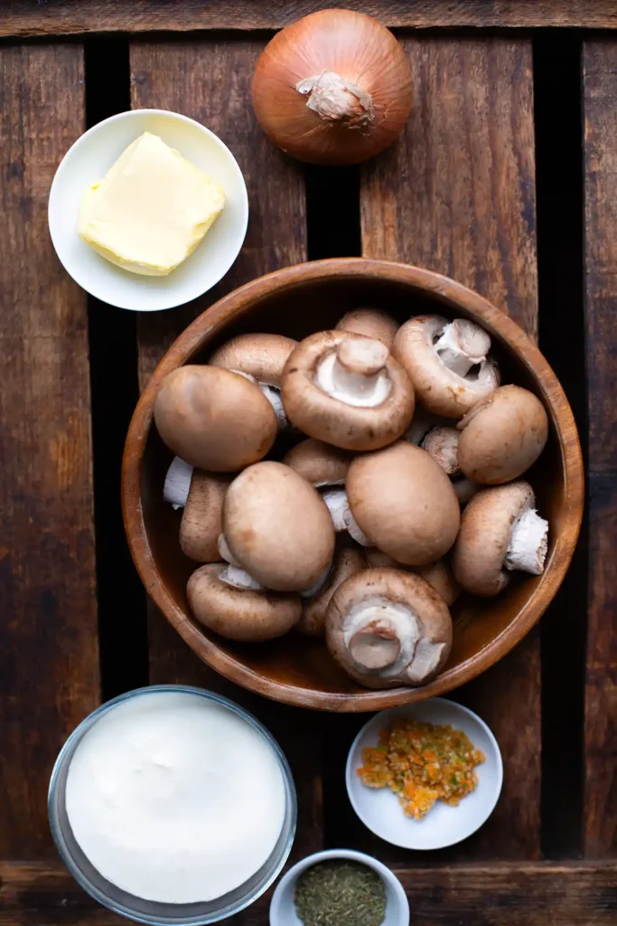 Zutaten für die Champignoncremesuppe. Zu sehen sind: eine weiße Zwiebel, braune Champignons, Butter, Gemüsebrühepaste, Thymian und Sahne. Die Zutaten stehen auf einem dunklen Holzuntergrund.