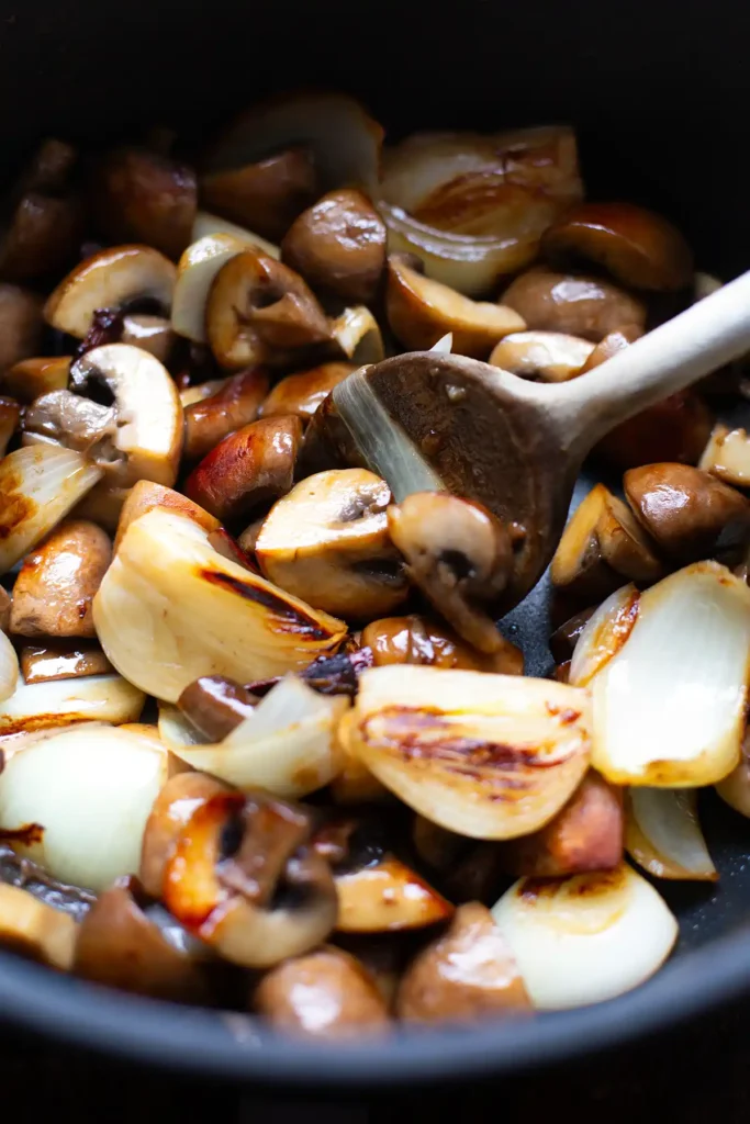 Klein geschnittene braune Champignons und weiße Zwiebel werden in einer schwarzen Pfanne angebraten und mit einem braunen Holzlöffel gewendet.