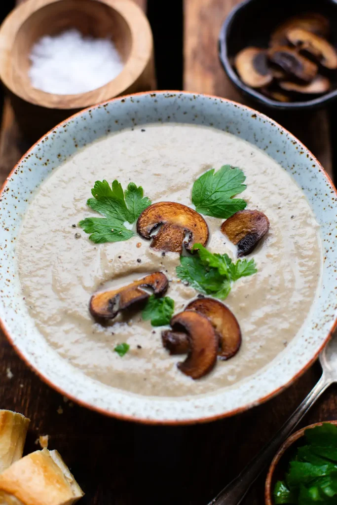 Fertige einfache Pilzsuppe in einer Schüssel, garniert mit glatter Petersilie und knusprig angebratenen Champignonscheiben. Daneben zwei weitere kleine Schüsseln: einmal mit Salz und einmal mit weiteren Scheiben von Champignons.