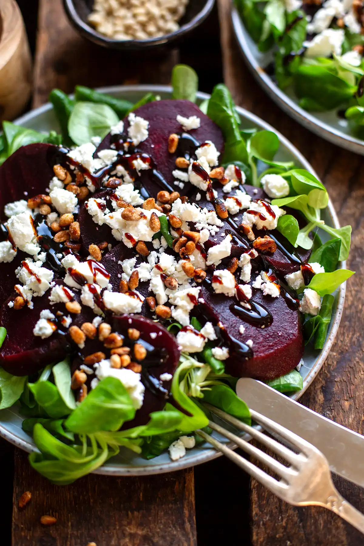 Fertiges Rote Bete Carpaccio auf einem grauen Teller. Angerichtet auf Feldsalat und garniert mit Balsamicocreme, zerbröseltem Feta und Pinienkernen. Daneben ist ein weiterer Teller und zwei Schälchen, einmal mit Pinienkernen drin zu sehen. Alles steht auf einem dunklen Holzuntergrund.