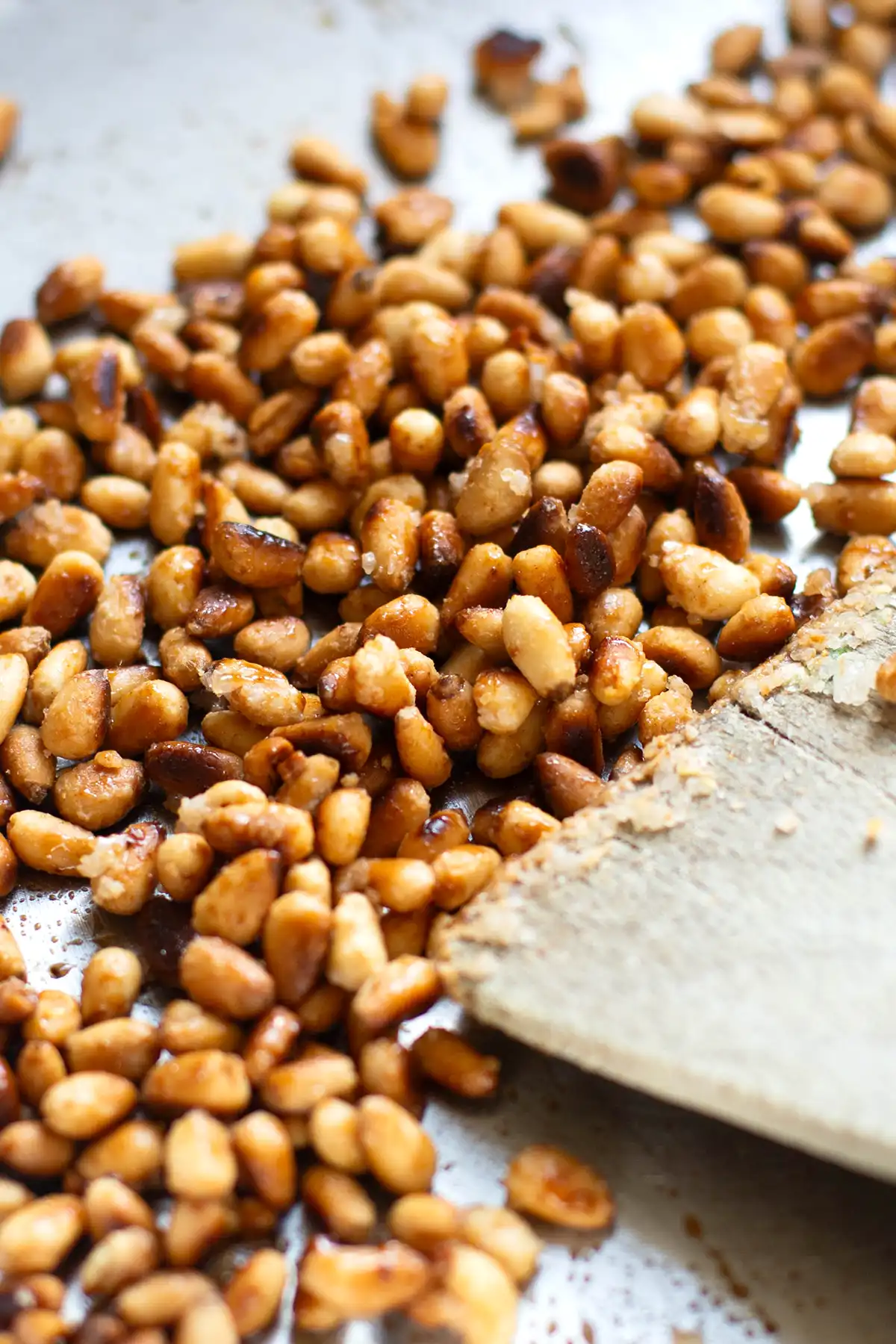 Geröstete Pinienkerne und mit Zucker karamellisierte Pinienkerne in einer silbernen Pfanne. Die Pinienkerne werden mit einem Holzpfannenwender gewendet.