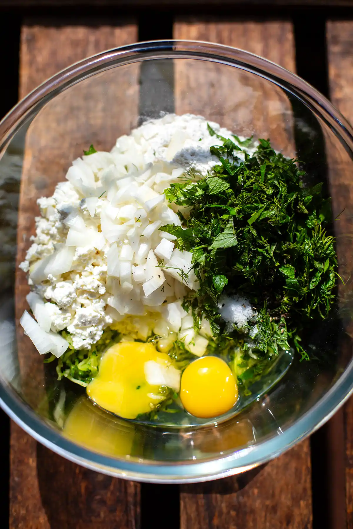 Alle Zutaten für die Zucchinipuffer in einer großen Glasschüssel kurz vor dem Verrühren. Zu sehen sind: klein gehackte weiße Zwiebel, gehackte Kräuter, Fetabrösel, zwei aufgeschlagene Eier und geraspelte Zucchini.