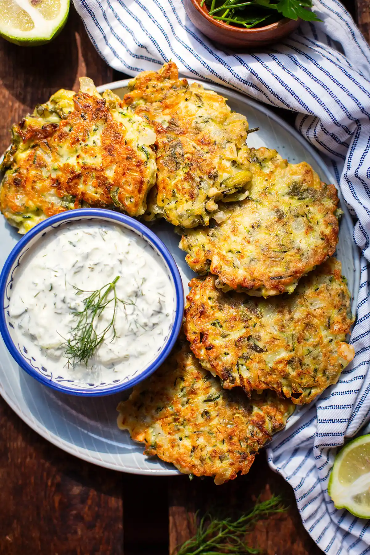 Fertige griechische Zucchinipuffer auf einem Teller. Darauf ebenfalls eine kleine blau-weiße Schüssel mit Tzatziki und Dill garniert. Der Teller steht auf einem dunklen Holzuntergrund. Neben dem Teller ein blau-weiß gestreiftes Geschirrtuch, eine kleine braune Schale mit Kräutern und eine halbe Limette.