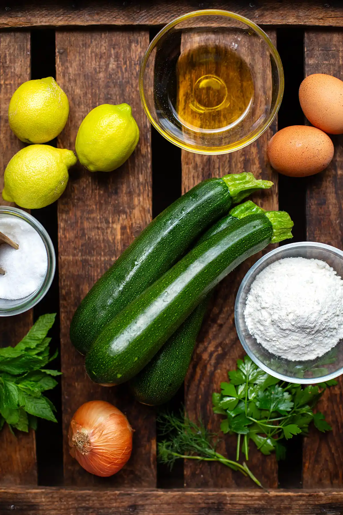 Die Zutaten für Zucchinipuffer einzeln auf einem dunklen Holzuntergrund. Zu sehen sind drei Zucchini, Mehl, Salz, Zitronen, Eier, Olivenöl, weiße Zwiebel und verschiedene Kräuter.