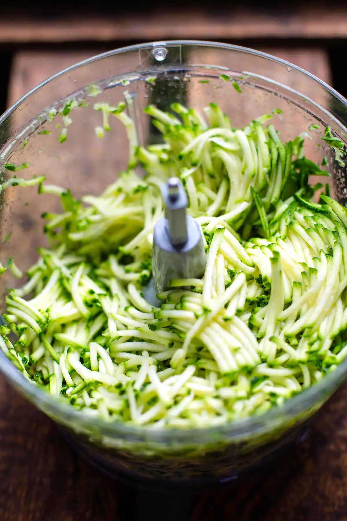 Geraspelte Zucchini für die Zucchinipuffer in einer Küchenmaschine. Das Gefäß steht auf einem dunklen Holzuntergrund.